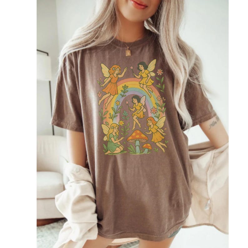 May include: Brown t-shirt featuring a whimsical design of fairies, a rainbow, and mushrooms. The fairies are depicted in various poses with colorful wings and dresses. The design is set against a backdrop of flowers and foliage.