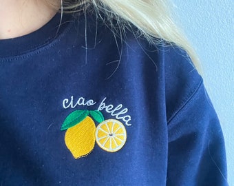 Könnte beinhalten: Ein marineblauer Sweatshirt mit einem gestickten Zitronenmotiv und dem Text "ciao bella" in Weiß.