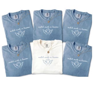 May include: Six t-shirts, five in light blue and one in white, are displayed. Each shirt has the phrase "match made in heaven" and an angel graphic. A gold-colored necklace with a pendant is on each shirt. The shirts are neatly folded.