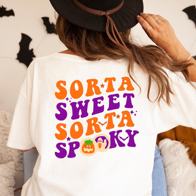 Pode incluir: Camiseta branca com o texto "Sorta Sweet Sorta Spooky" em laranja e roxo em fonte retr&ocirc;. O texto &eacute; decorado com estrelas e morcegos. Um gr&aacute;fico de ab&oacute;bora e fantasma est&aacute; abaixo do texto.