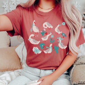 May include: A maroon t-shirt with a graphic print of geese in various poses. The geese are surrounded by flowers, strawberries, and other details.