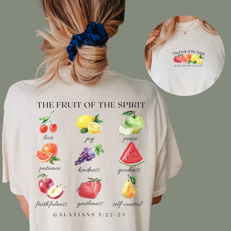 May include: A beige t-shirt featuring a graphic of fruit and the text "THE FRUIT OF THE SPIRIT" and "GALATIANS 5:22-23". The fruit includes a cherry, lemon, apple, orange, grapes, watermelon, apple, strawberry, and pear.