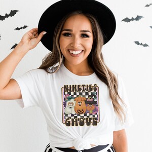 May include: White t-shirt featuring a graphic of three cartoon characters in Halloween costumes. The characters are a ghost, a pumpkin, and a witch. The graphic is surrounded by a chequered pattern and the text "Nineties Ghoul".