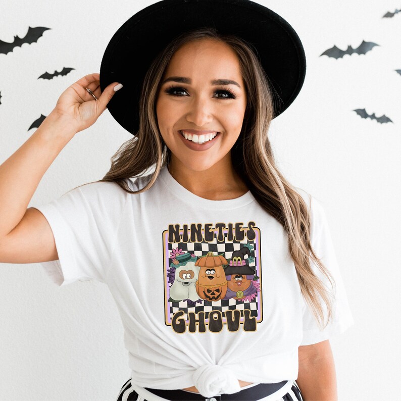 May include: White t-shirt featuring a graphic of three cartoon characters in Halloween costumes. The characters are a ghost, a pumpkin, and a witch. The graphic is surrounded by a chequered pattern and the text "Nineties Ghoul".