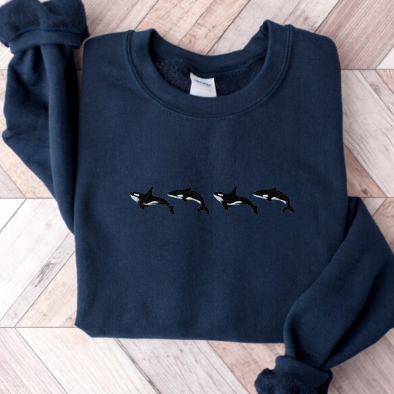 May include: Navy blue sweatshirt featuring an embroidered design of four black and white orcas swimming horizontally across the chest. The sweatshirt has a crew neck and long sleeves. The orcas are detailed with white bellies and black dorsal fins.