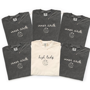 May include: A collection of six t-shirts, primarily in dark gray and cream colors. Each shirt features the words "inner circle" or "high lady" in script, along with a crescent moon and star graphic. A delicate necklace is draped on each shirt.