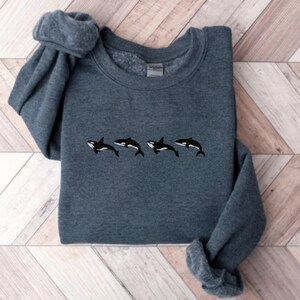 May include: A dark teal sweatshirt featuring four embroidered orca whales in black and white. The whales are horizontally aligned across the chest of the sweatshirt. The sleeves are folded over.