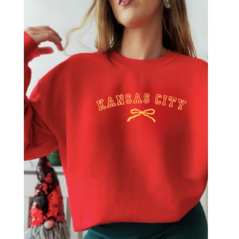 May include: Red sweatshirt with a yellow embroidered design that says "Kansas City" with a yellow bow underneath.