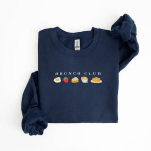 Pu&ograve; includere: Felpa blu navy con la scritta "BRUNCH CLUB" sopra una fila di icone ricamate di cibi per il brunch. Le icone includono un uovo, una fragola, un rotolo alla cannella, caff&egrave; e pancake. La felpa &egrave; realizzata in un materiale morbido.
