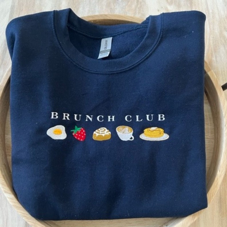 Pu&ograve; includere: Felpa blu navy con la scritta ricamata "BRUNCH CLUB" e illustrazioni di cibi per la colazione: un uovo fritto, una fragola, un rotolo alla cannella, una tazza di caff&egrave; e pancake. La felpa &egrave; piegata.