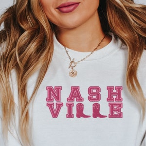 May include: White sweatshirt with a pink "NASHVILLE" graphic with two pink cowboy boots. The sweatshirt has a gold chain necklace with a gold charm.