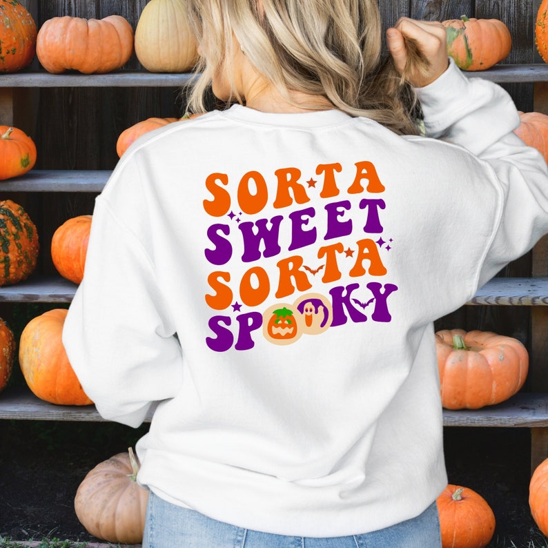 May include: White sweatshirt with the words "Sorta Sweet Sorta Spooky" in orange and purple. The design includes a jack-o'-lantern, a ghost, and a bat. The background features pumpkins on wooden shelves.