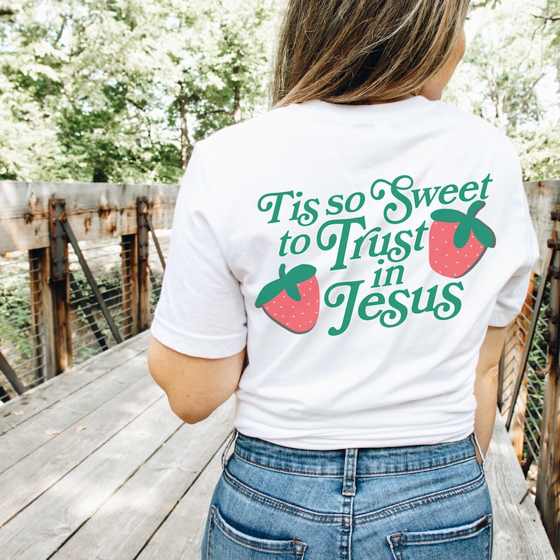 May include: White t-shirt with a green and red graphic that says "Tis so Sweet to Trust in Jesus" with two strawberries on either side of the text.