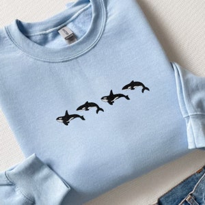 May include: A light blue sweatshirt featuring a line of four embroidered black and white orca whales. The sweatshirt has a crew neck and long sleeves. The garment is made of a soft material.