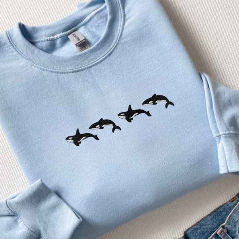 May include: A light blue sweatshirt featuring a line of four embroidered black and white orca whales. The sweatshirt has a crew neck and long sleeves. The garment is made of a soft material.