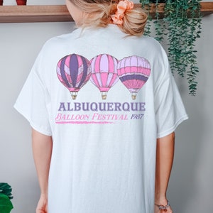Op de afbeelding: Wit T-shirt met een roze en paarse afbeelding van drie luchtballonnen. De tekst "Albuquerque Balloon Festival 1987" is onder de ballonnen gedrukt.