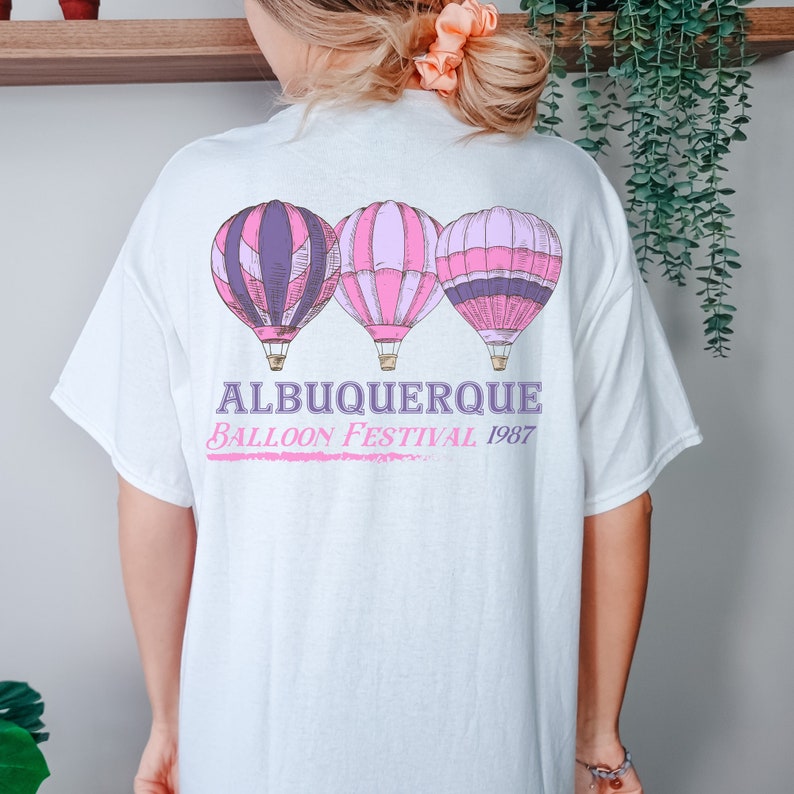 Op de afbeelding: Wit T-shirt met een roze en paarse afbeelding van drie luchtballonnen. De tekst "Albuquerque Balloon Festival 1987" is onder de ballonnen gedrukt.