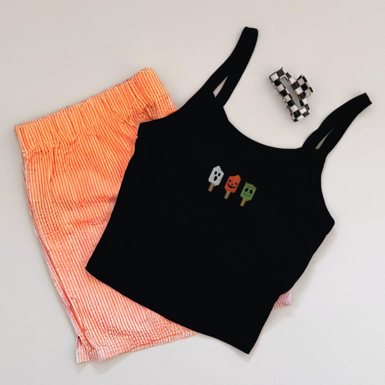 May include: A black tank top with three embroidered Halloween-themed popsicle designs, paired with orange striped shorts and a checkered hair clip. The shorts and tank top are made of a soft material.