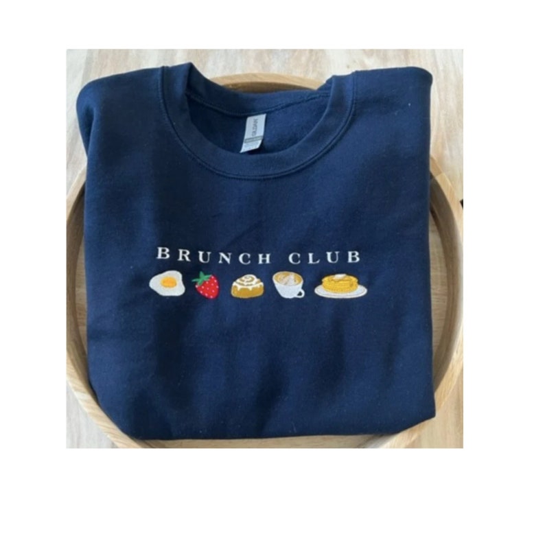 Pu&ograve; includere: Felpa blu navy con la scritta ricamata "BRUNCH CLUB" sopra illustrazioni ricamate di cibi per la colazione: un uovo, una fragola, un rotolo alla cannella, caff&egrave; e pancake. La felpa &egrave; piegata in un vassoio di legno.