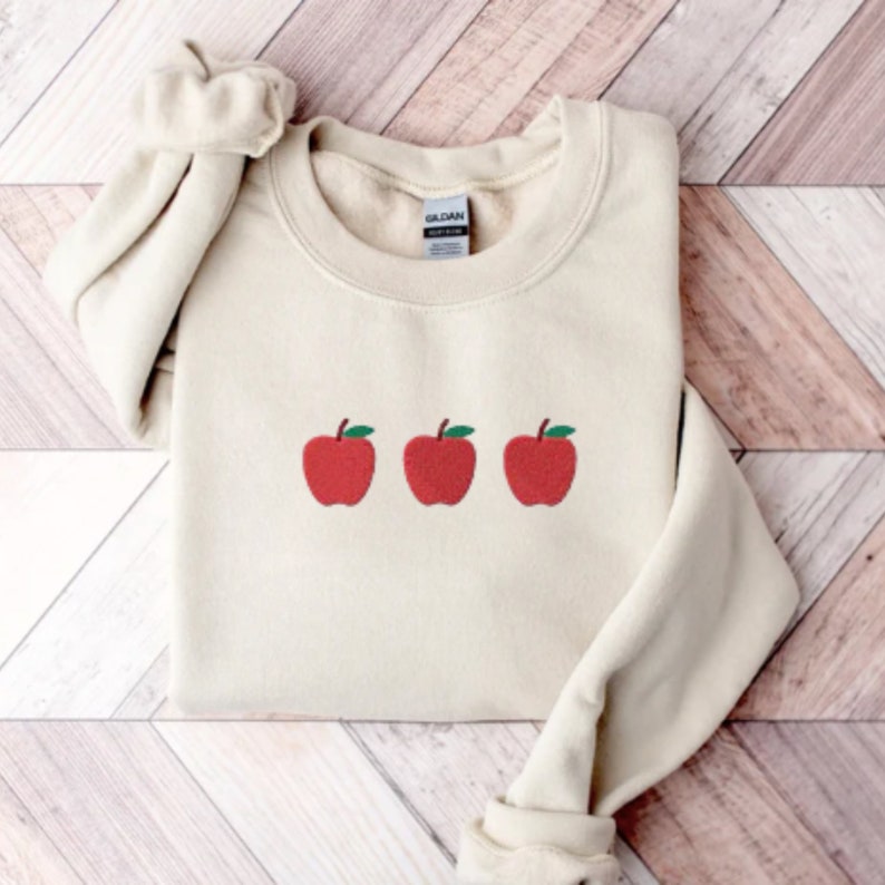 May include: A cream-colored sweatshirt with three red apples embroidered on the front. The apples have green leaves and stems.
