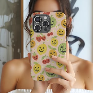 May include: A phone case with a Halloween-themed pattern. The design features smiling jack-o'-lanterns, strawberries, lemons, cherries, and watermelons on a light pink background. The case has a glossy finish and a camera cutout.
