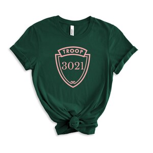 May include: A dark green t-shirt with a pink crest design. The crest reads "TROOP" at the top and "3021" in the center. The shirt is made of a soft material and has a crew neck.