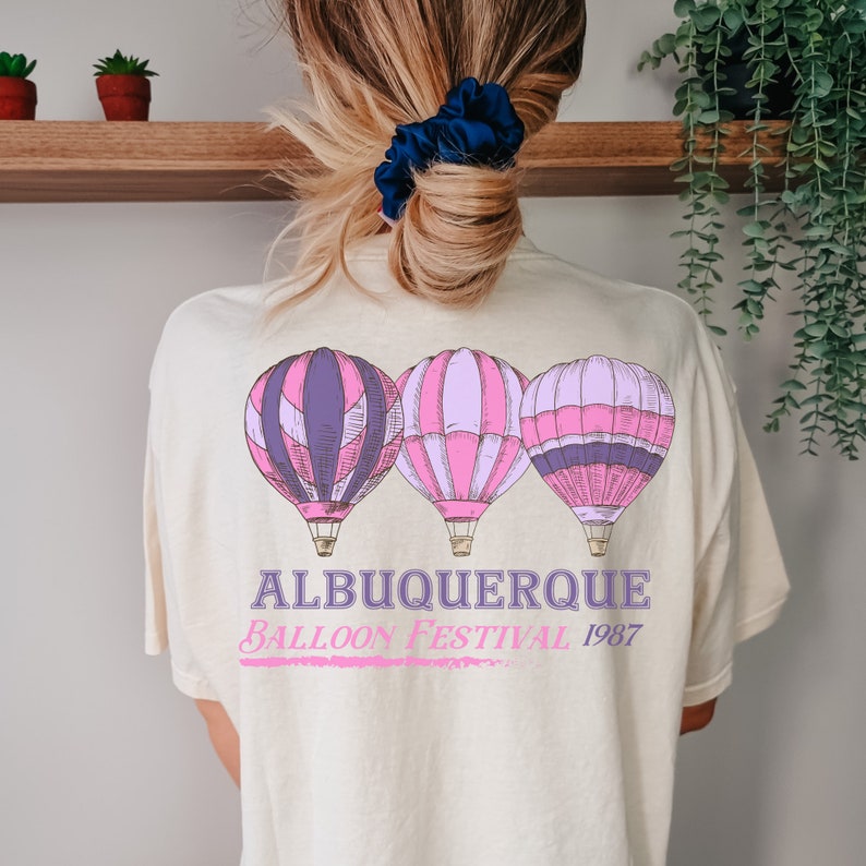 Op de afbeelding: Een cr&egrave;mekleurig T-shirt met een afbeelding van drie luchtballonnen in roze, paars en wit. De tekst "Albuquerque Balloon Festival 1987" is onder de ballonnen gedrukt.