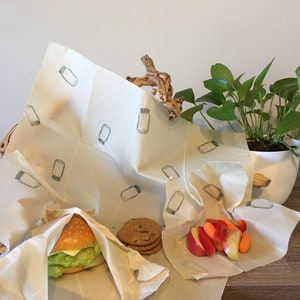 Puede incluir: Envoltorios de alimentos de cera de abeja con un patrón de tarro, utilizados para envolver una hamburguesa, galletas y rodajas de manzanas y zanahorias. Los envoltorios reutilizables son de color beige y están sobre una superficie de madera.