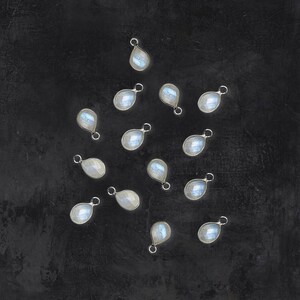 May include: A collection of 15 silver teardrop-shaped charms with iridescent white moonstone gemstones. The charms are scattered on a black background.