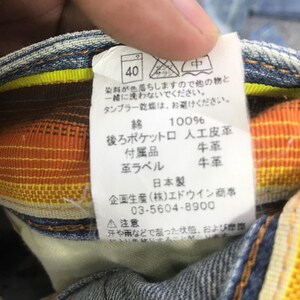 K&ouml;nnte beinhalten: Nahaufnahme eines Denim-Kleidungsst&uuml;cketiketts mit japanischem Text. Das Etikett zeigt Waschanweisungen, Stoffzusammensetzung und Markeninformationen. Das Etikett ist wei&szlig; mit schwarzem Text und einer kleinen Grafik. Das Kleidungsst&uuml;ck hat orange und gelbe Akzente.