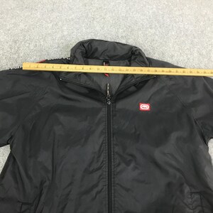 May include: A black zip-up jacket with a high collar and a red Ecko Unltd logo patch on the chest. A yellow wooden ruler is placed horizontally across the top, showing a width measurement of approximately 26 inches from shoulder to shoulder.