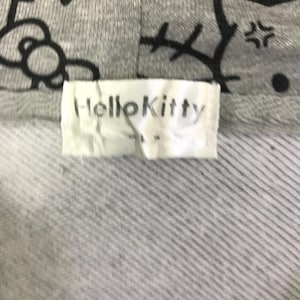 Może przedstawiać: Zbliżenie szarej bluzy z kapturem z motywem Hello Kitty. Etykieta głosi "HelloKitty" czarnymi literami. Bluza z kapturem ma teksturowany, prążkowany wz&oacute;r.