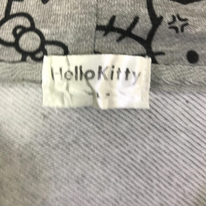 Może przedstawiać: Zbliżenie szarej bluzy z kapturem z motywem Hello Kitty. Etykieta głosi "HelloKitty" czarnymi literami. Bluza z kapturem ma teksturowany, prążkowany wz&oacute;r.