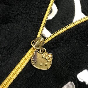 Pu&ograve; includere: Una giacca in pile nera e bianca con una cerniera dorata e un ciondolo Hello Kitty dorato.