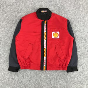 May include: A red and black jacket with a zipper closure and a Shell logo on the front. The jacket has a black and yellow stripe down the front zipper.