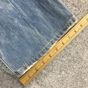 K&ouml;nnte beinhalten: Nahaufnahme von hellblauem Denim-Stoff mit ausgefranstem Rand. Ein Holzlineal liegt diagonal &uuml;ber dem Stoff und zeigt Messungen in Zentimetern an. Der Denim hat sichtbare N&auml;hte und ein abgenutztes Aussehen.