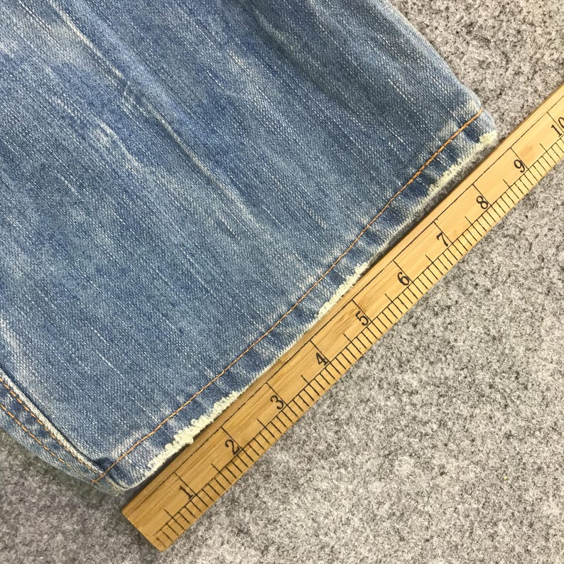 K&ouml;nnte beinhalten: Nahaufnahme von hellblauem Denim-Stoff mit ausgefranstem Rand. Ein Holzlineal liegt diagonal &uuml;ber dem Stoff und zeigt Messungen in Zentimetern an. Der Denim hat sichtbare N&auml;hte und ein abgenutztes Aussehen.