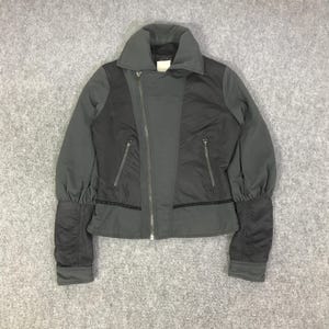 Könnte beinhalten: Dunkelgraue und schwarze Jacke mit hohem Kragen und diagonalem Reißverschluss. Die Jacke hat eine taillierte Silhouette mit Puffärmeln und zwei Reißverschlusstaschen. Die Jacke besteht aus einer Materialkombination mit mattem Finish.