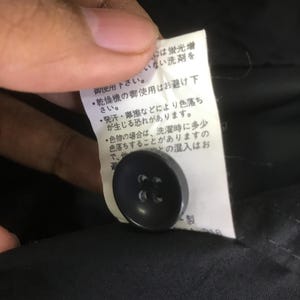 May include: A black four-hole button attached to a white fabric care label featuring Japanese text instructions. The label is positioned against a black garment background, providing clear visibility of the button and the printed care information.