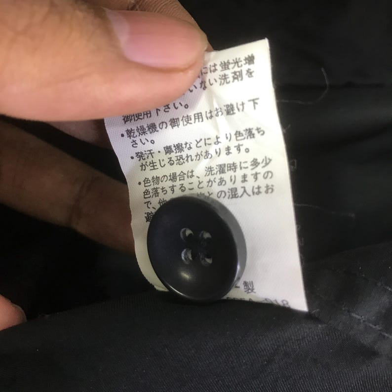 May include: A black four-hole button attached to a white fabric care label featuring Japanese text instructions. The label is positioned against a black garment background, providing clear visibility of the button and the printed care information.