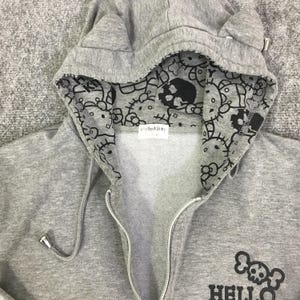 Może przedstawiać: Szara bluza z kapturem zapinana na zamek z kapturem w kształcie uszu kota. Wnętrze kaptura jest wzorzyste z czarnymi grafikami Hello Kitty i czaszek. Bluza ma logo Hello Kitty z czaszką z przodu.
