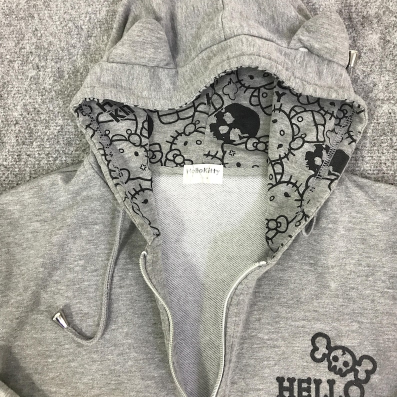 Może przedstawiać: Szara bluza z kapturem zapinana na zamek z kapturem w kształcie uszu kota. Wnętrze kaptura jest wzorzyste z czarnymi grafikami Hello Kitty i czaszek. Bluza ma logo Hello Kitty z czaszką z przodu.