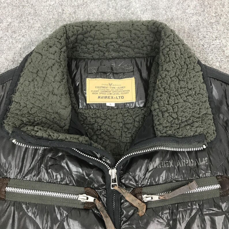 Może przedstawiać: Czarna pikowana kurtka z kołnierzem z ciemnozielonego sztucznego futra. Kurtka posiada zapięcie na zamek błyskawiczny i etykietę z napisem "EQUIPMENT-TYPE-AVIREX FLIGHT GARMENT SPECIFICATION HIGH SPEED LOW LEVEL FLIGHT AVIREX LTD".