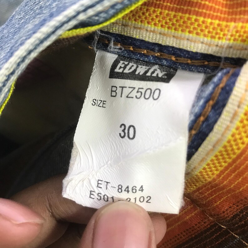 K&ouml;nnte beinhalten: Nahaufnahme eines wei&szlig;en Etiketts mit dem Markennamen "Edwin", dem Text "BTZ500" und "SIZE". Die Zahl "30" ist auf dem Etikett aufgedruckt. Das Etikett ist an einer Jeans befestigt.