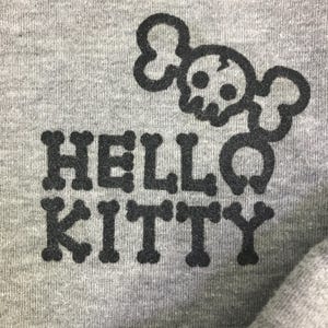 Może przedstawiać: Zbliżenie szarej tkaniny z czarną grafiką. Grafika przedstawia słowa "HELLO KITTY" w czcionce przypominającej kości, z motywem czaszki i skrzyżowanych piszczeli nad "HELLO". Tkanina ma lekko teksturowany wygląd.