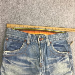 K&ouml;nnte beinhalten: Nahaufnahme einer hellblauen Jeans mit Used-Details. Merkmale sind orangefarbene N&auml;hte, ein Knopfverschluss und ein gestreifter Bund. Die Jeans hat Vordertaschen und eine verwaschene Optik.