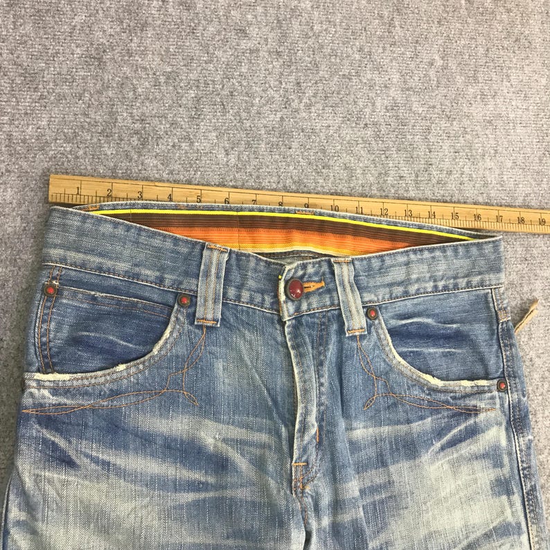K&ouml;nnte beinhalten: Nahaufnahme einer hellblauen Jeans mit Used-Details. Merkmale sind orangefarbene N&auml;hte, ein Knopfverschluss und ein gestreifter Bund. Die Jeans hat Vordertaschen und eine verwaschene Optik.
