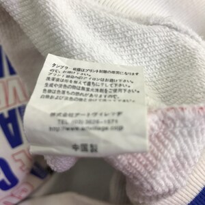 以下が含まれることがあります： 白いスウェットシャツのクローズアップ。タグには日本語のテキストと「Made in China」の文字があります。スウェットシャツには青、赤、白のアクセントがあります。