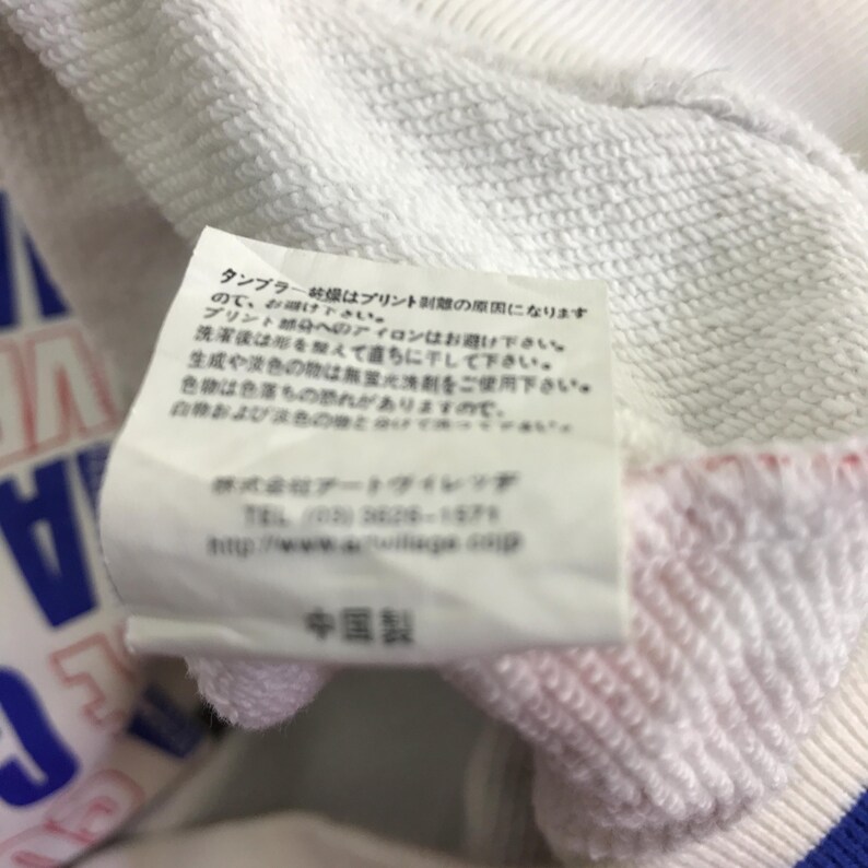 以下が含まれることがあります： 白いスウェットシャツのクローズアップ。タグには日本語のテキストと「Made in China」の文字があります。スウェットシャツには青、赤、白のアクセントがあります。
