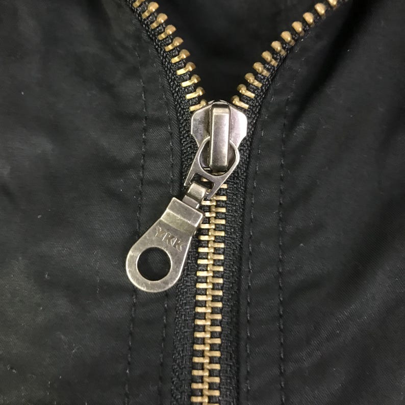 May include: A black fabric garment featuring a metal zipper closure with brass-toned teeth and a silver-toned pull tab. The pull tab is circular with a rectangular attachment point and features the embossed text YKK on its surface.